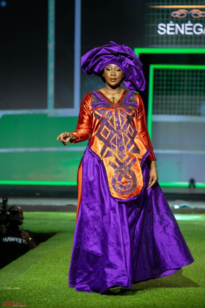 Modèle en tenue violette et orange devant le panneau Sénégal à l'Afrik Fashion Week