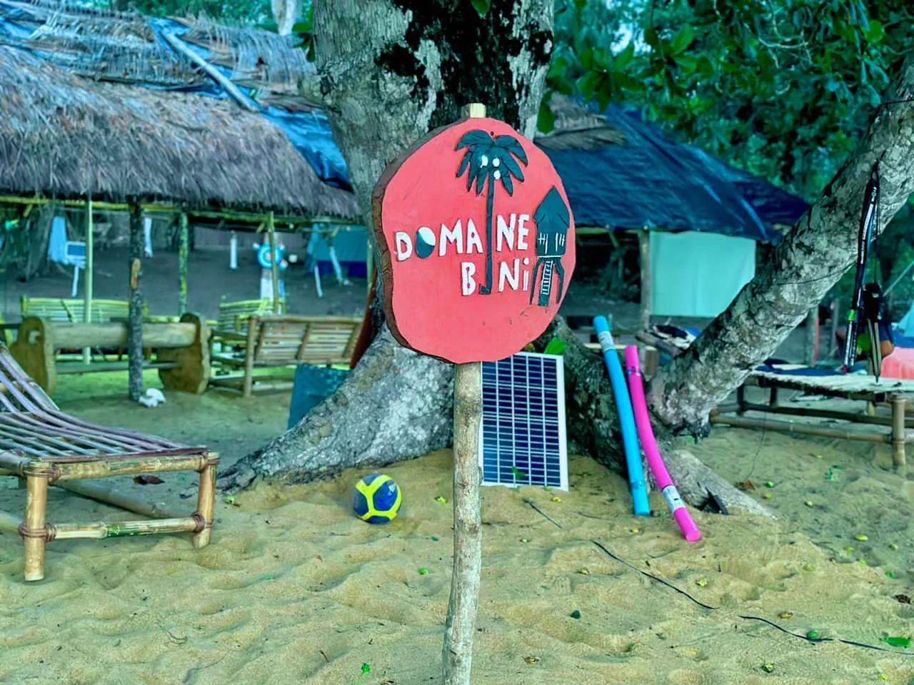 The hand painted sign " Domaine Bini" in front of the bamboo mats and deckchairs of the camp